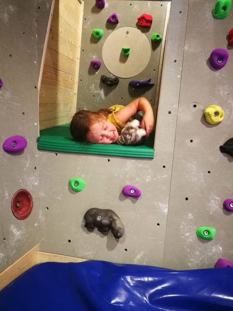 Ein Kind liegt entspannt in einer Kletterwand mit bunten Griffen und umarmt ein Spielzeug.