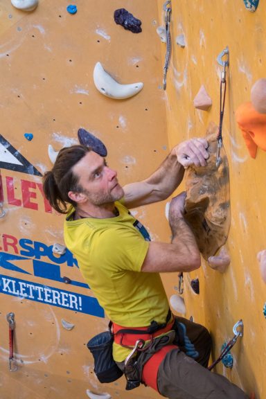 Ein Kletterer in gelbem Shirt greift nach einem Griff an einer Kletterwand.