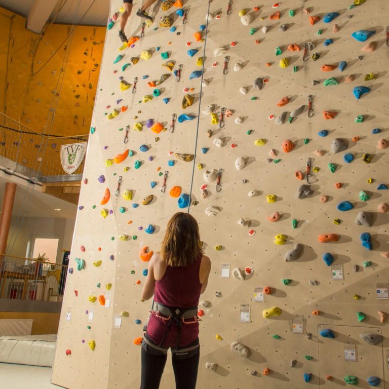 Eine Person steht vor einer bunten Kletterwand in einer Kletterhalle.