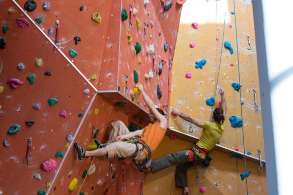 Zwei Personen beim Klettern an einer bunten Kletterwand in einer Sporthalle.