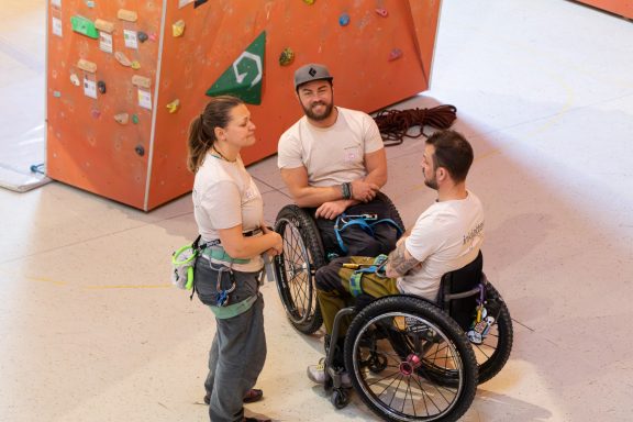 Drei Personen, zwei im Rollstuhl, sprechen in einer Kletterhalle.