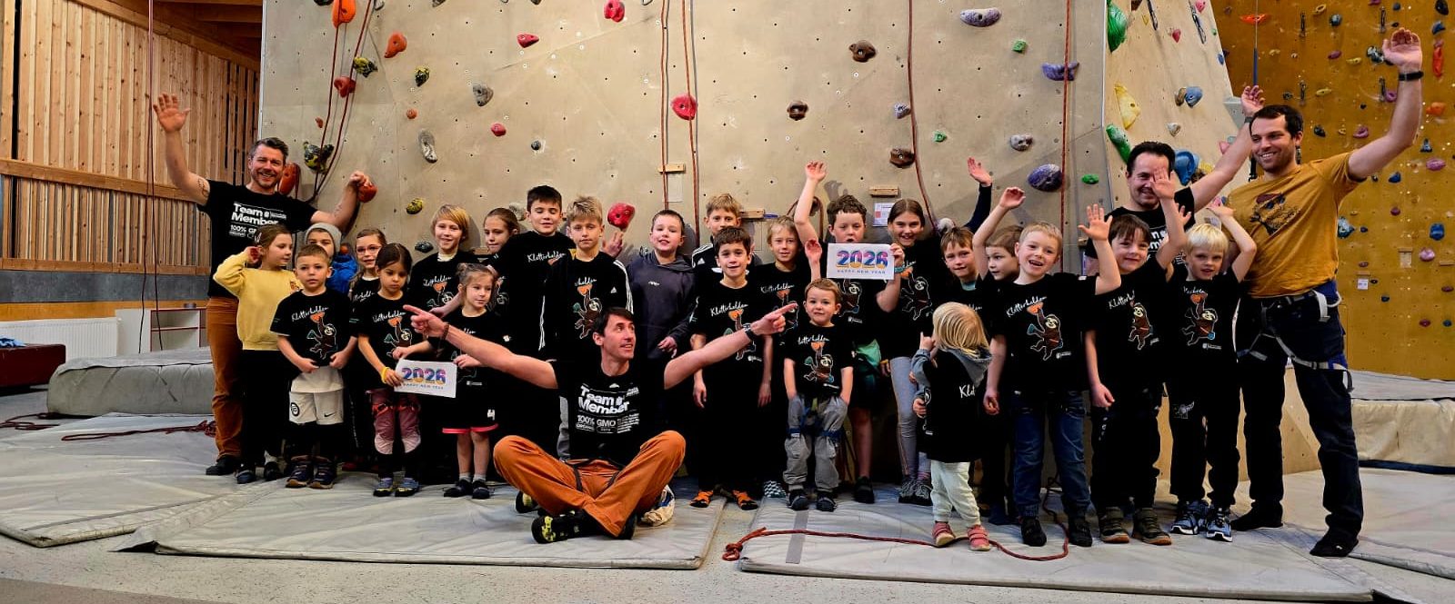 Nachwuchs Kletterer der Kletterhalle Fürstenfeld Gruppe von Kindern und Erwachsenen in Kletterausrüstung, lächelnd vor einer Kletterwand.