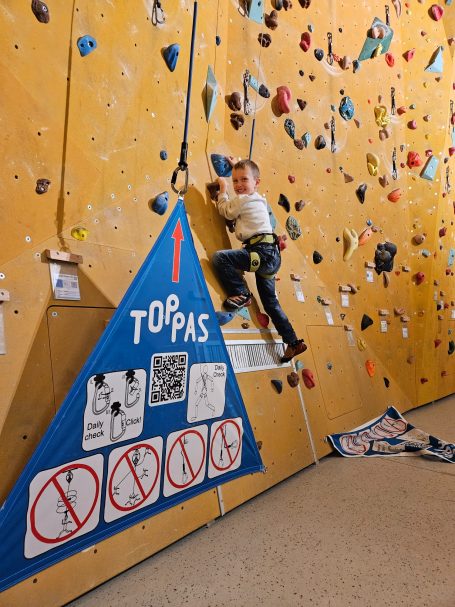 Junger Kletterer an einer Kletterwand mit Hinweisschildern darunter.